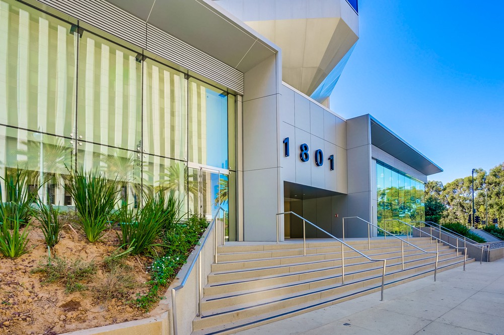 Barrister Suites Office Lease A modern office building with large glass windows, silver panels, and the address number 1801 displayed above a set of stairs with metal railings. There are plants in a landscaped area beside the entrance.