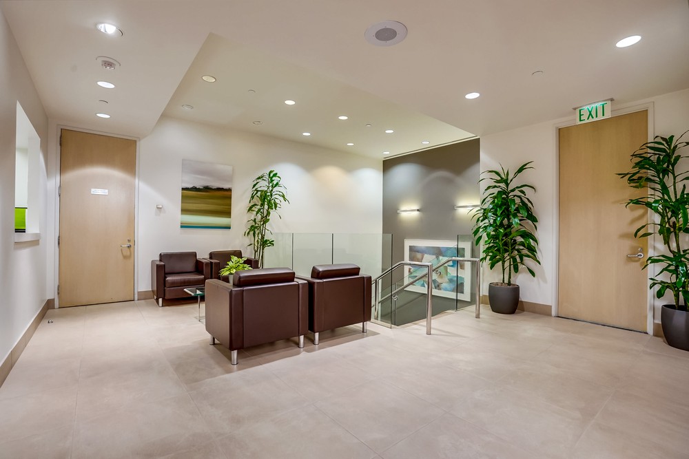 Barrister Suites Office Lease A modern office lobby with brown leather chairs, a small table, potted plants, beige walls and flooring, artwork on the wall, and doors on both sides. Stairs lead down from the seating area. An exit sign is above one door.