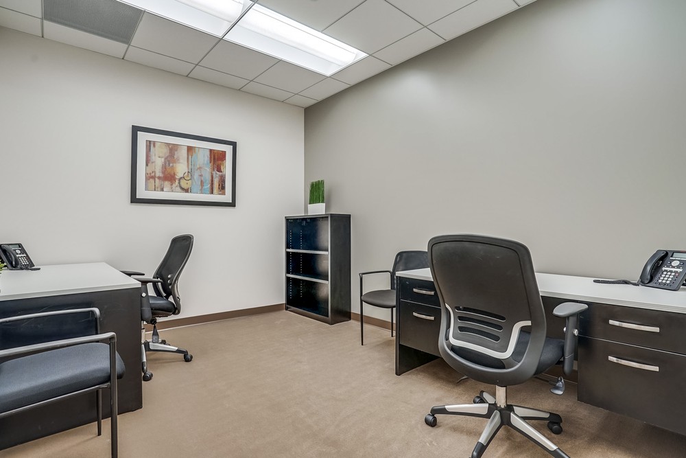 Barrister Suites Office Lease A small office with three desks, black chairs, telephones, a black shelf with a green plant, and abstract art on a gray wall. The room is well-lit with overhead fluorescent lights.