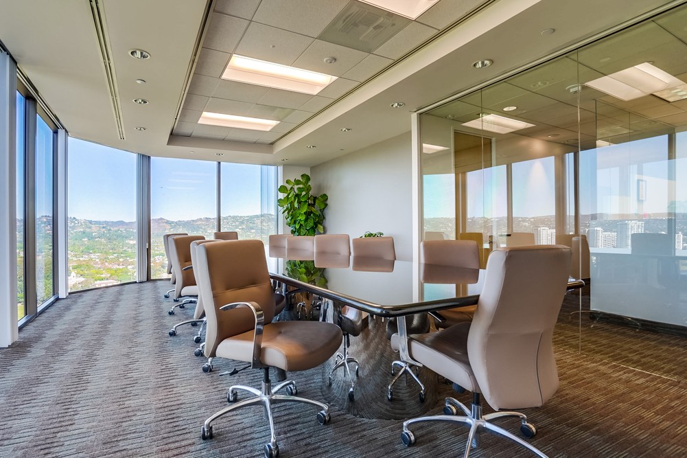 Barrister Suites Office Lease Modern conference room with a long glass table surrounded by beige swivel chairs, large floor-to-ceiling windows with a cityscape view, and a potted plant in the corner. The room is well-lit and has neutral decor.