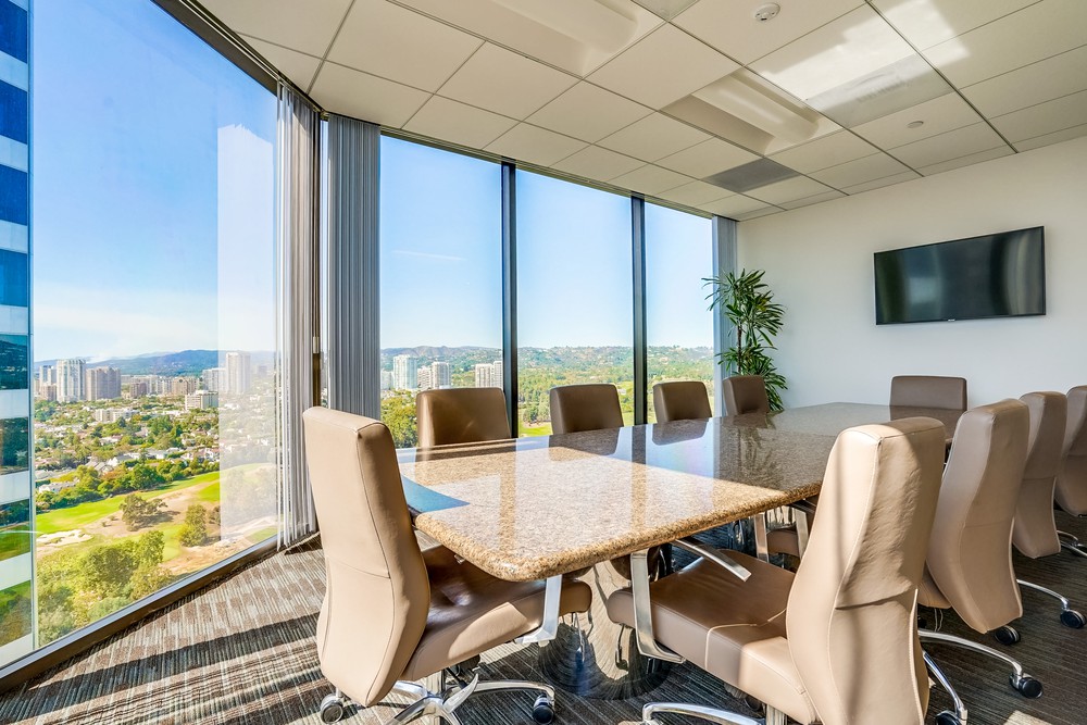 Barrister Suites Office Lease A modern conference room with a large marble table, tan leather chairs, floor-to-ceiling windows showing a cityscape and greenery, a wall-mounted TV, and a tall plant in the corner.