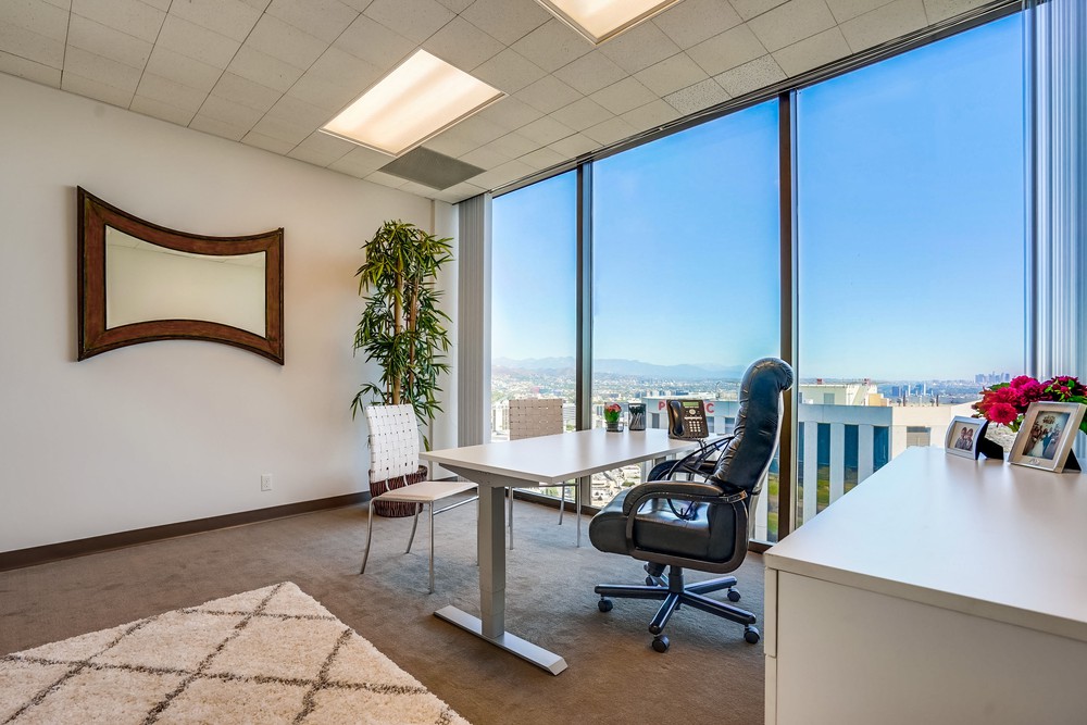 Barrister Suites Office Lease A modern office with large floor-to-ceiling windows, a desk with a black chair, a white guest chair, a potted plant, a decorative mirror, and city views under a clear blue sky.