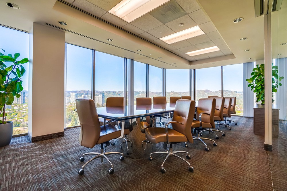 Barrister Suites Office Lease A modern conference room with a long table surrounded by brown chairs, large floor-to-ceiling windows offering a city view, and green potted plants near the windows. The room is well-lit and spacious.