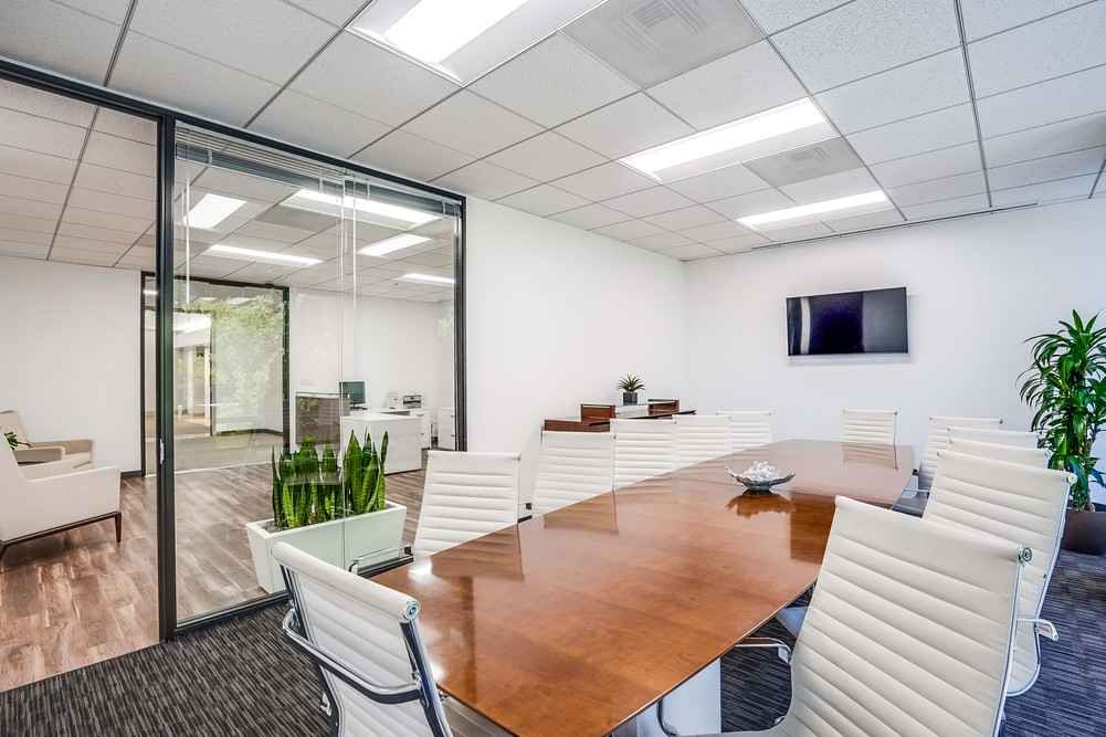 Barrister Suites Office Lease Modern conference room with a long wooden table, white chairs, a wall-mounted TV, potted plants, and glass walls overlooking a hallway and adjacent office space. The room is bright with overhead lighting.