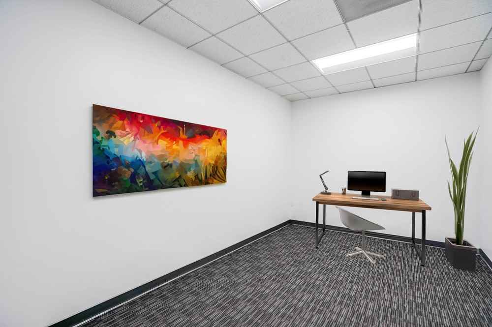 Barrister Suites Office Lease A minimalist office with a wooden desk, computer, lamp, and chair. A colorful abstract painting hangs on the white wall, and a tall plant is in the corner. The floor is carpeted with a striped pattern.