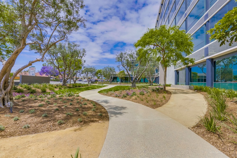 Barrister Suites Office Lease A winding sidewalk runs through a landscaped garden with trees and shrubs beside a modern glass building under a partly cloudy sky.
