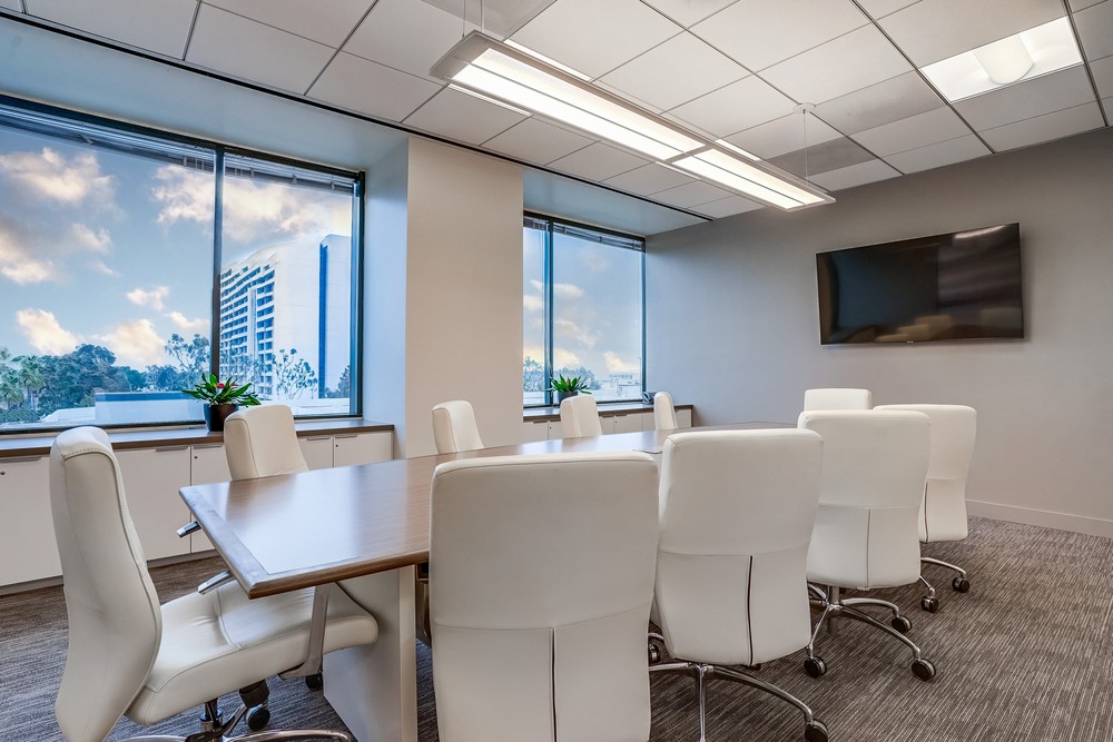 Barrister Suites Office Lease Modern conference room with a wooden table surrounded by eight white office chairs, large windows with city views, two potted plants, overhead lights, and a wall-mounted flat screen TV.