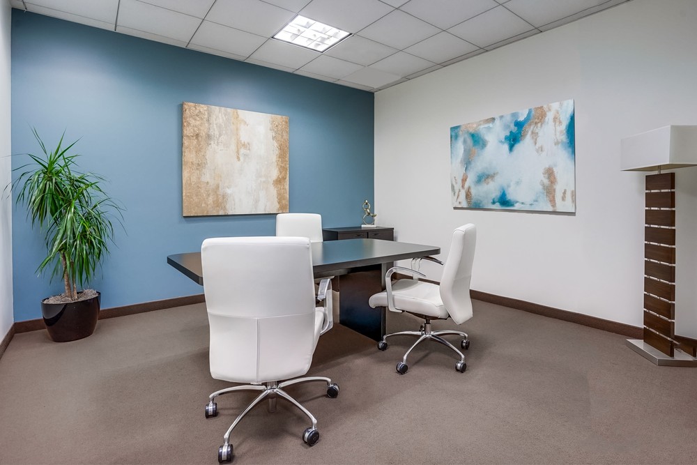 Barrister Suites Office Lease Modern office with a blue accent wall, two white chairs at a black desk, a potted plant, abstract paintings on the walls, and a tall floor lamp in the corner. The room is well-lit with overhead lights.