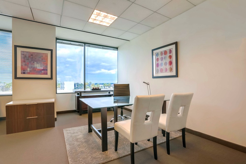 Barrister Suites Office Lease Modern office with a desk, black chair, two white chairs, large window with city view, a side cabinet, and abstract art on the walls. The space is bright with natural light and minimal decor.