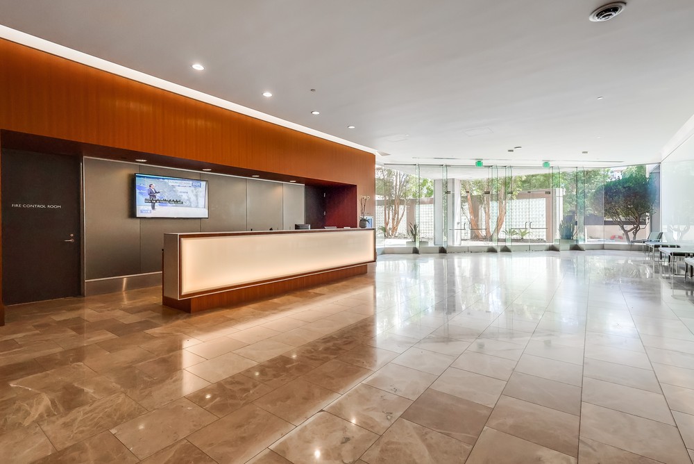Barrister Suites Office Lease Spacious, modern lobby with a shiny marble floor, a long reception desk with warm lighting, large glass windows, and trees visible outside. A wall-mounted screen and several doors are on the left.