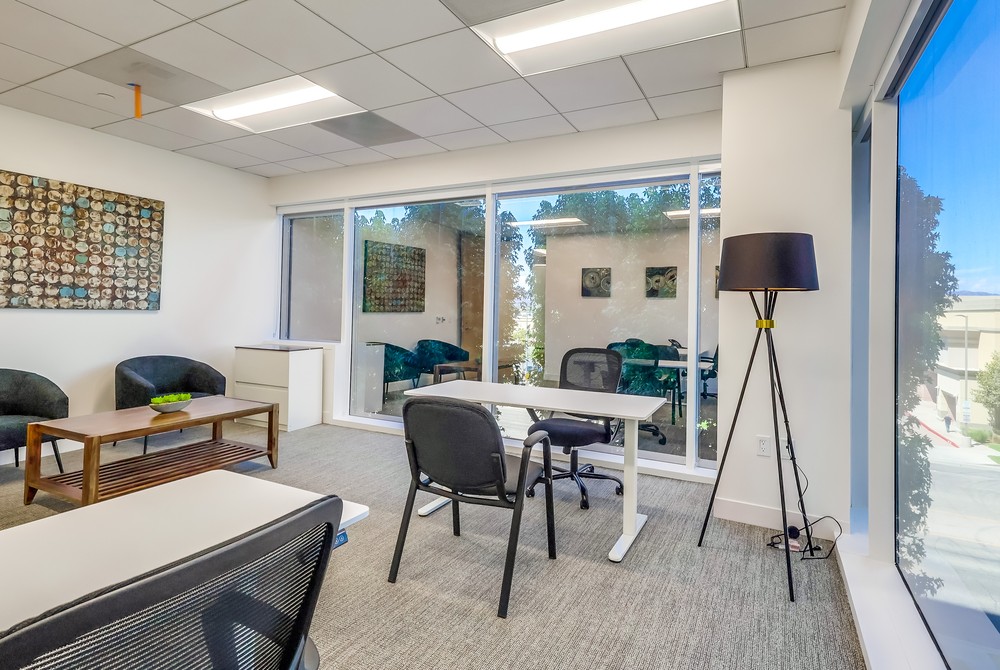 Barrister Suites Office Lease Bright modern office with large windows, white walls, a desk and chair, a floor lamp, coffee table, and two armchairs. Art decor hangs on the wall and trees are visible outside.