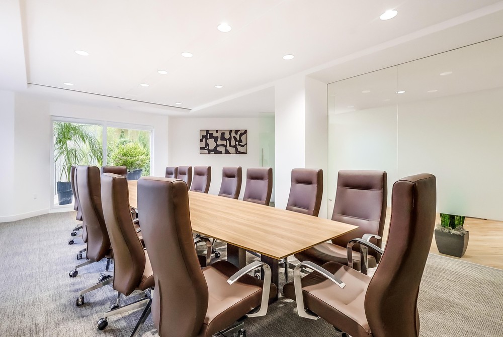 Barrister Suites Office Lease A modern conference room with a long wooden table surrounded by high-back brown chairs, large windows with greenery outside, gray carpet, and abstract art on the white wall.