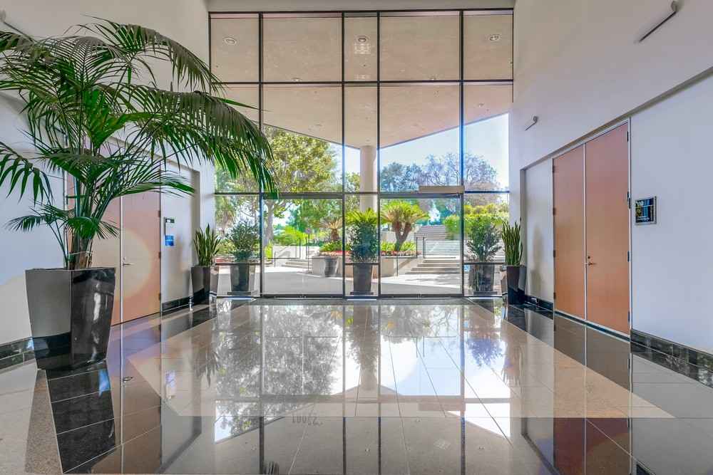 Barrister Suites Office Lease Modern building lobby with large glass windows, tall potted plants, reflective tiled floor, and double wooden doors. Sunlight streams in, revealing outdoor greenery and a patio area beyond the windows.
