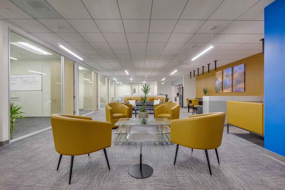 Barrister Suites Office Lease Modern office lounge with yellow chairs around glass tables, gray carpet, glass-walled offices, potted plants, and abstract artwork on a wood-paneled wall. The space is brightly lit with ceiling lights.