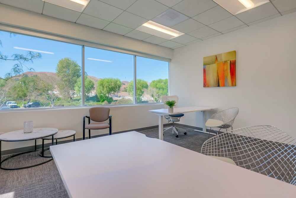 Barrister Suites Office Lease Bright, modern office with large windows, three desks, chairs, small round tables, a potted plant, and colorful abstract art on the wall. Trees and blue sky are visible outside.