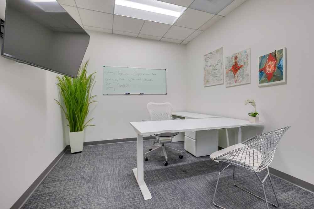 Barrister Suites Office Lease A modern office with a white desk, two chairs, a wall-mounted TV, a whiteboard with writing, green plant, small orchid, and abstract artwork on the walls. The room has gray carpeting and bright overhead lighting.
