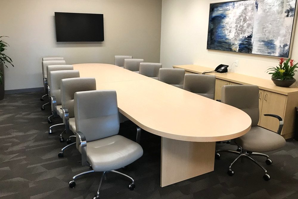 Barrister Suites Office Lease A modern conference room with a long oval table surrounded by twelve gray chairs, a wall-mounted TV, a large abstract painting, a phone, and potted plants on a cabinet.