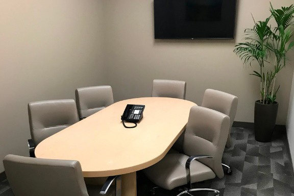 Barrister Suites Office Lease A small conference room with a light wood oval table, six beige chairs, a phone on the table, a wall-mounted TV, a potted plant in the corner, and carpeted flooring.