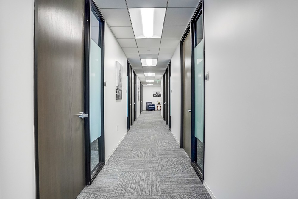 Barrister Suites Office Lease A clean, modern office hallway with gray carpet, white walls, and doors with glass panels on both sides, leading to a distant open area with chairs. Bright ceiling lights illuminate the corridor.