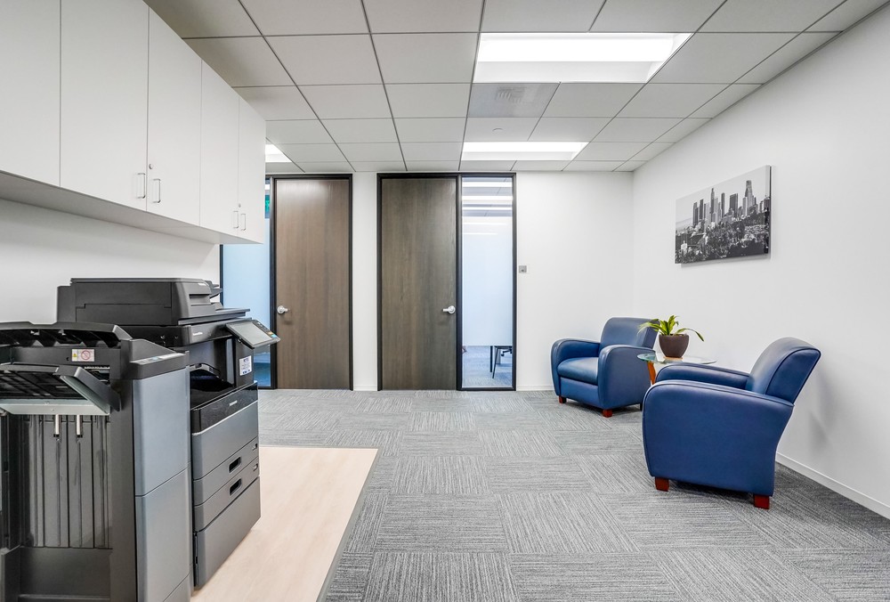 Barrister Suites Office Lease Modern office space with two blue armchairs, a small table with a plant, a copier/printer, storage cabinets, and two closed wooden doors; a black-and-white cityscape photo hangs on the white wall.