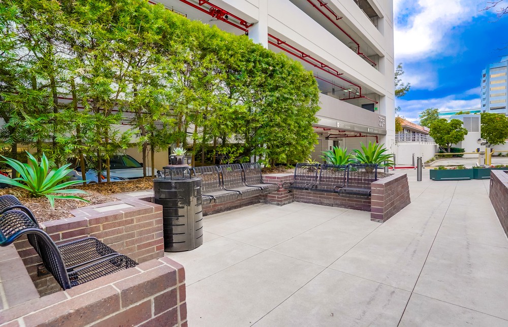 Barrister Suites Office Lease Outdoor seating area with black metal benches, brick planters, green plants, and a trash can beside a multi-level parking garage, under a partly cloudy sky.