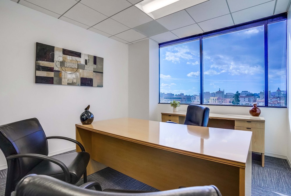 Barrister Suites Office Lease A modern office with a wooden desk, two black chairs, abstract wall art, and a large window showing a cityscape under a bright blue sky with clouds. A small plant and vase sit on the desk by the window.