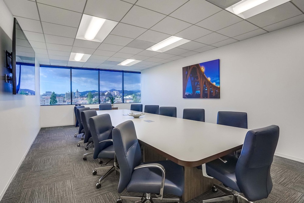 Barrister Suites Office Lease Modern conference room with a long table surrounded by blue chairs, large windows offering a city view, white walls, overhead lights, and a framed photo of a bridge on the wall.