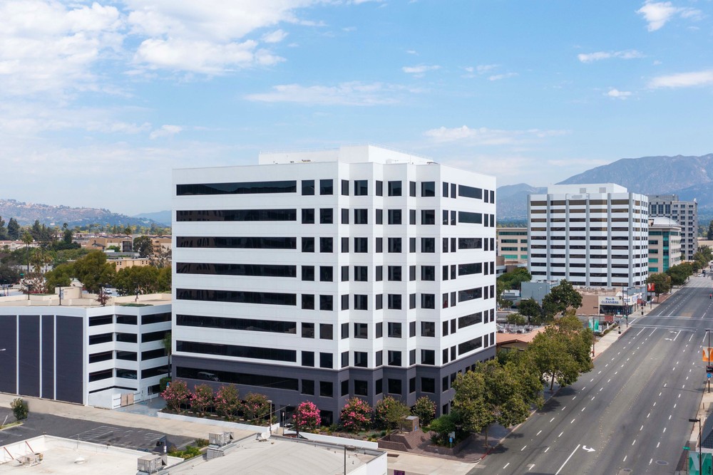 Barrister Suites Office Lease A modern, mid-rise office building with white and black horizontal stripes stands beside a wide, empty street lined with trees. Other office buildings and mountains are visible in the background under a partly cloudy sky.