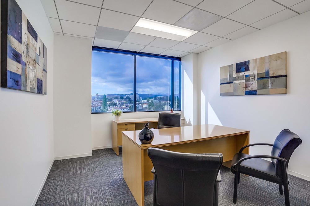 Barrister Suites Office Lease Modern office with a wooden desk, two black chairs, abstract art on white walls, gray carpet, and a large window showing a cityscape and cloudy sky outside.