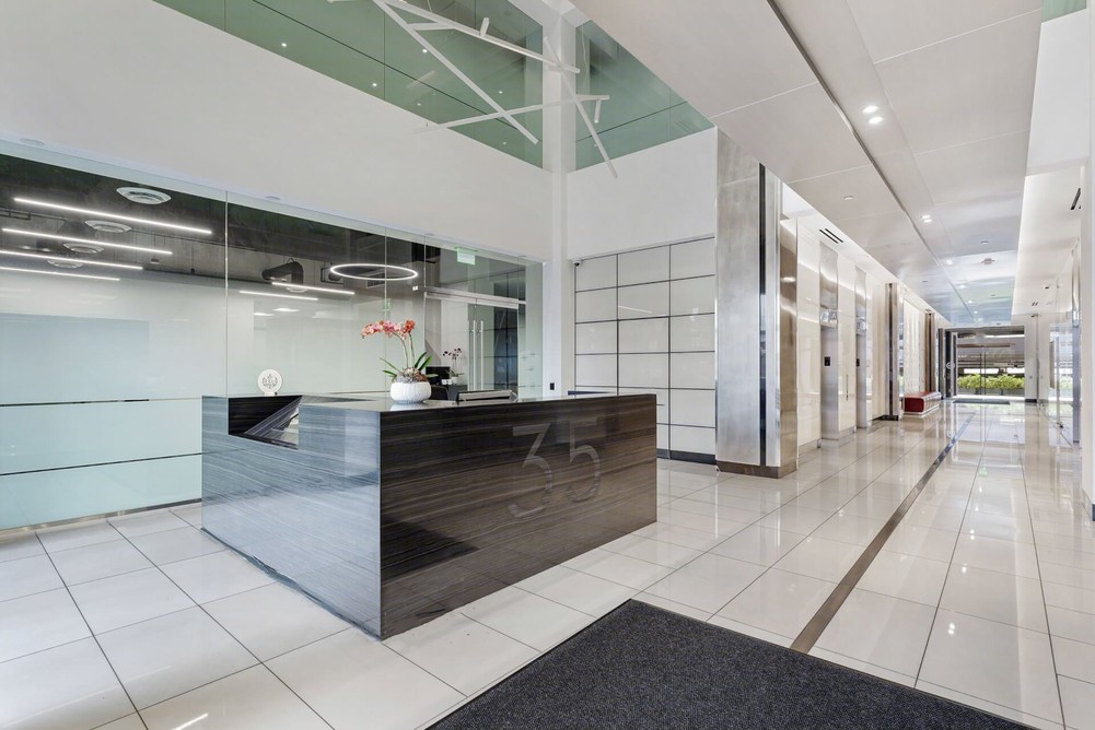 Barrister Suites Office Lease Modern office lobby with a glossy black reception desk, glass walls, tiled floors, white walls, and ceiling lights. A potted orchid sits on the desk, and there is a hallway leading to glass doors and greenery outside.