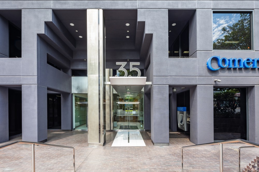 Barrister Suites Office Lease A modern building entrance with dark gray walls and large glass doors. The number 35 is displayed above the entrance. Part of a Comerica Bank sign and an outdoor ATM are visible on the right side.