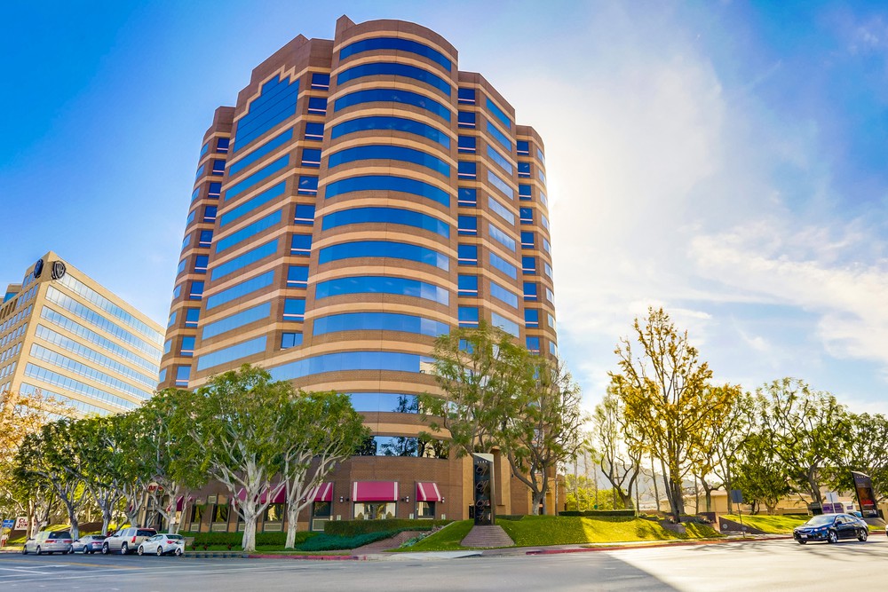 Barrister Suites Office Lease A modern, multi-story office building with curved edges and many windows, surrounded by trees and a clear blue sky. The sun is shining brightly, casting shadows on the street and sidewalk in front of the building.