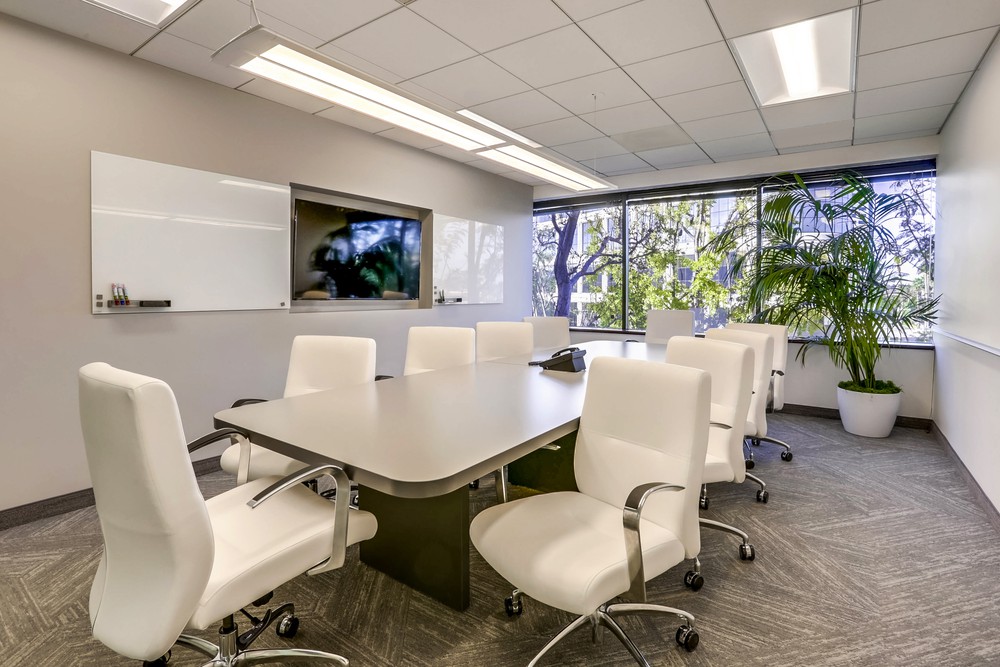 Barrister Suites Office Lease Modern conference room with a rectangular table surrounded by six white chairs, a whiteboard, wall-mounted TV, large windows with trees visible outside, and a potted plant in the corner.