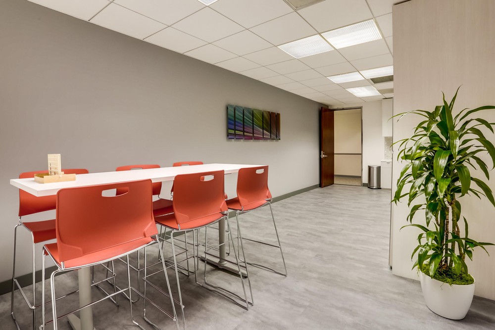 Barrister Suites Office Lease Modern office break room with a tall white table and six red chairs, wall art, gray walls, and a potted plant in the corner. The room has a tiled ceiling and bright overhead lighting.