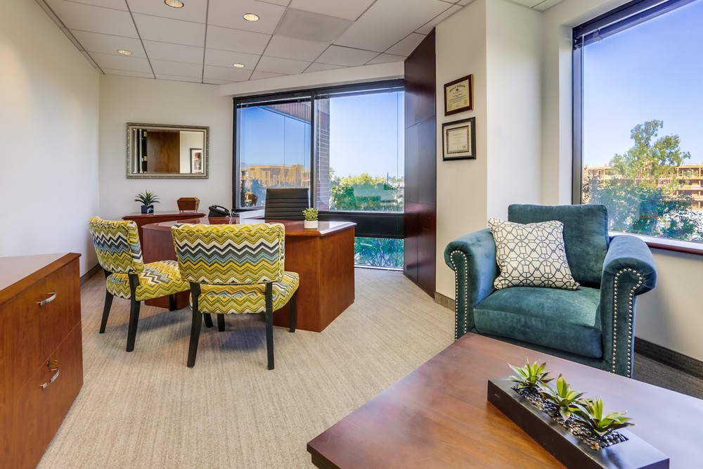 Barrister Suites Office Lease A modern office with a wooden desk, two patterned yellow chairs, a teal armchair with a decorative pillow, a window with a city view, wall art, and certificates. A small plant sits on a coffee table in the foreground.