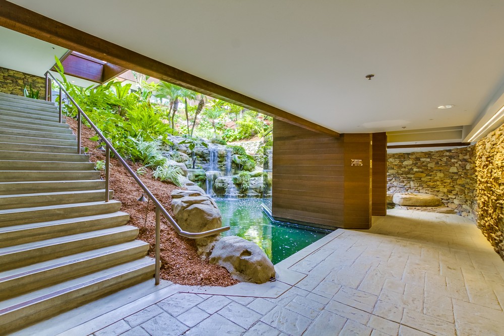 Barrister Suites Office Lease A modern indoor space with stone walls and flooring features a staircase beside a large window, revealing a lush garden with a waterfall flowing into a pond. Natural light fills the area.