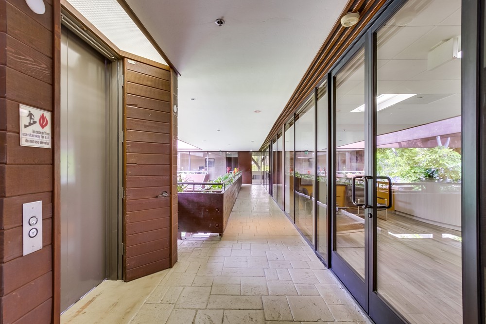 Barrister Suites Office Lease A modern hallway with tiled flooring, brown wooden walls, an elevator on the left, large glass windows and doors on the right, and green plants visible outside.