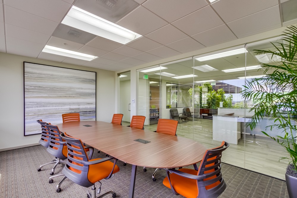 Barrister Suites Office Lease A modern conference room with a wooden table, seven orange chairs, a large abstract painting on the wall, and glass walls overlooking a bright office area with plants.