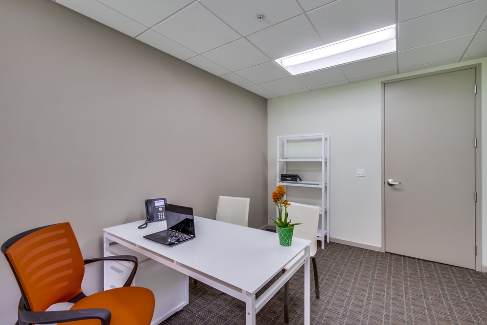 Barrister Suites Office Lease A small office with a white desk, orange and white chairs, a laptop, a phone, and a potted plant. There is a white shelving unit and a closed beige door in the background. Fluorescent lights brighten the space.