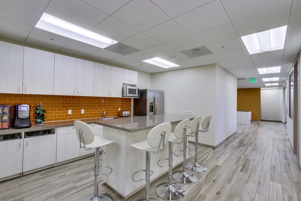 Barrister Suites Office Lease Modern office kitchen with white cabinets, orange tile backsplash, stainless steel appliances, a long island with four white bar stools, and wood-style flooring. Bright overhead lighting and a hallway are visible.