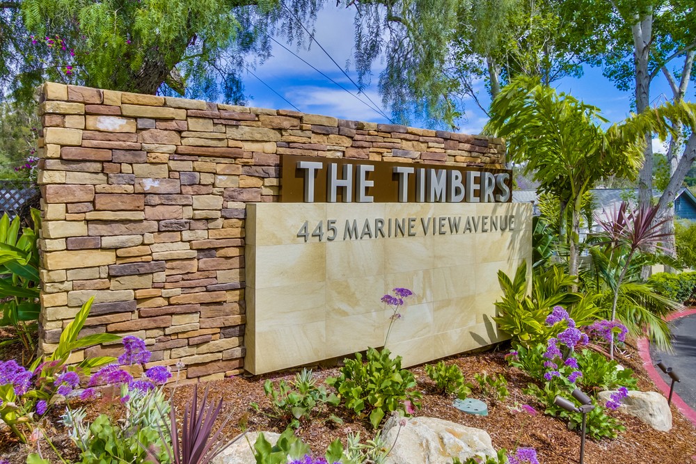 Barrister Suites Office Lease A stone sign surrounded by greenery and purple flowers reads THE TIMBERS and 445 MARINE VIEW AVENUE. Trees and a blue sky are visible in the background.