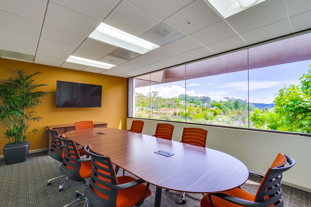Barrister Suites Office Lease Modern conference room with an oval table, orange chairs, a wall-mounted TV, a large potted plant, and a wide window offering a scenic view of greenery and hills outside.