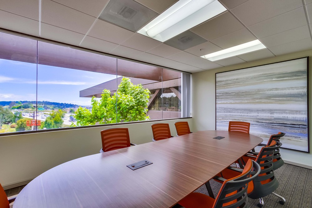 Barrister Suites Office Lease A modern conference room with a long wooden table, orange chairs, large window with a scenic outdoor view, and a large abstract painting on the wall.