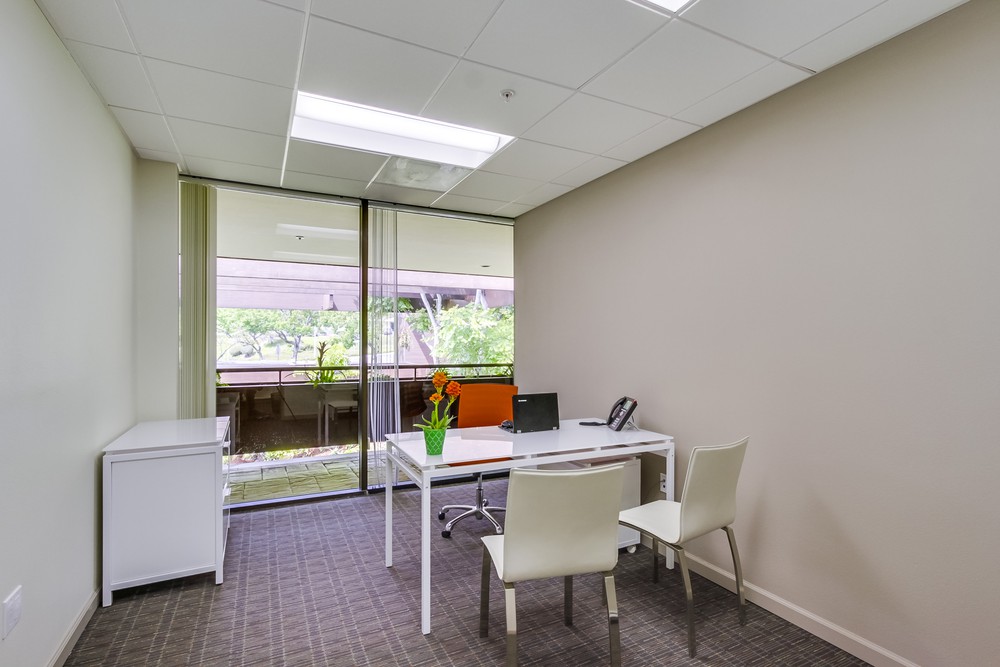 Barrister Suites Office Lease A modern, minimal office with a white desk, two white chairs, a laptop, a phone, a green plant, and a cabinet. Large windows overlook an outdoor area with greenery and natural light fills the room.