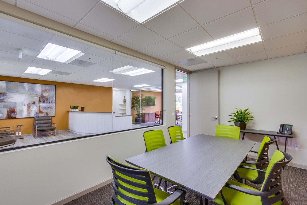 Barrister Suites Office Lease A modern office meeting room with a gray table, six green chairs, a potted plant, and a phone. A glass wall separates the room from a reception area with a painting, chairs, and orange accent wall.