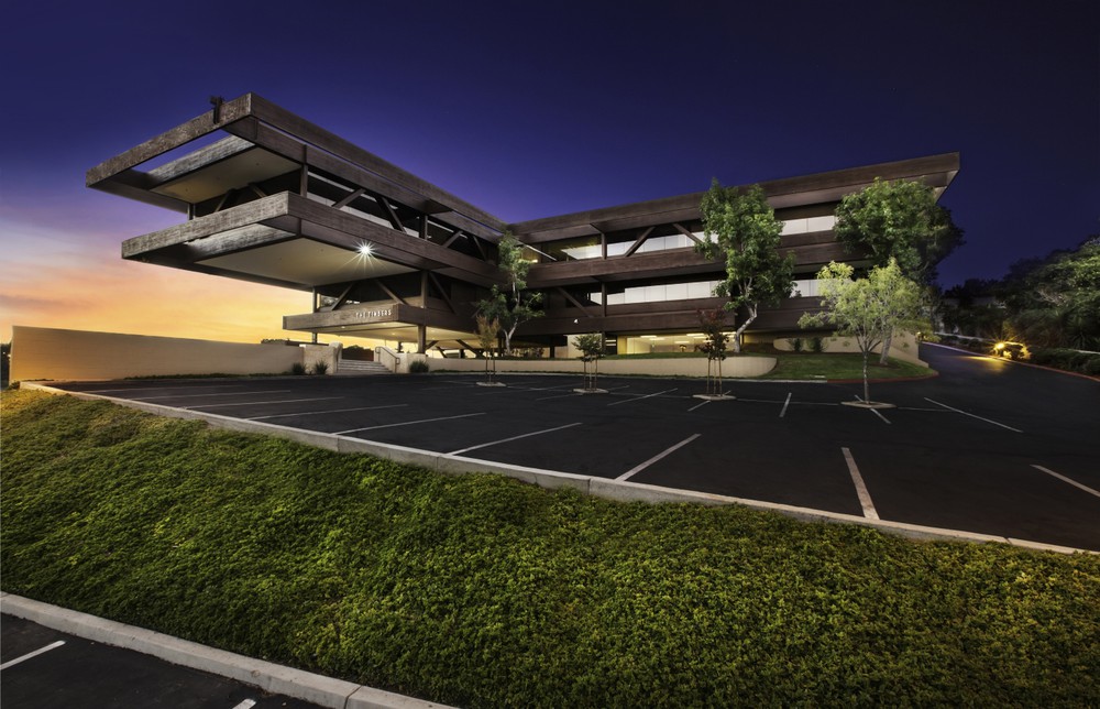 Barrister Suites Office Lease A modern, angular office building with large overhangs and glass windows is shown at dusk, with lights on inside. The parking lot in front is empty, and there are trees and green landscaping in the foreground.