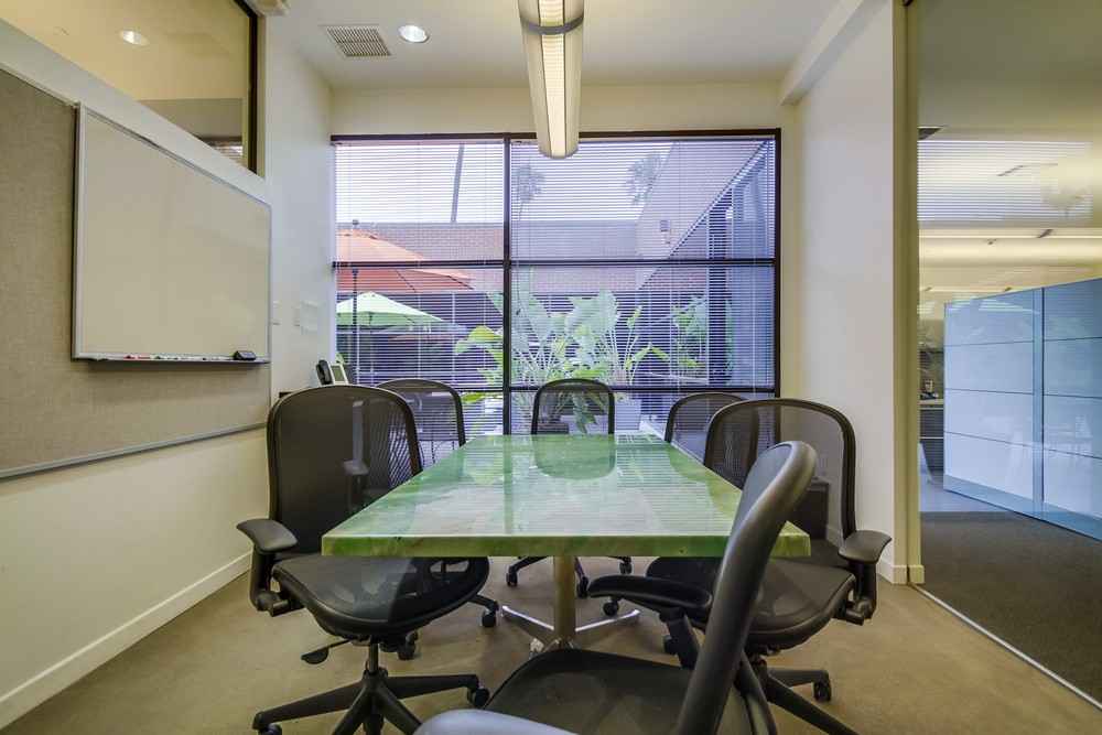Barrister Suites Office Lease A small conference room with a green rectangular table surrounded by six black office chairs, large windows with blinds, and a view of plants and an orange umbrella outside.