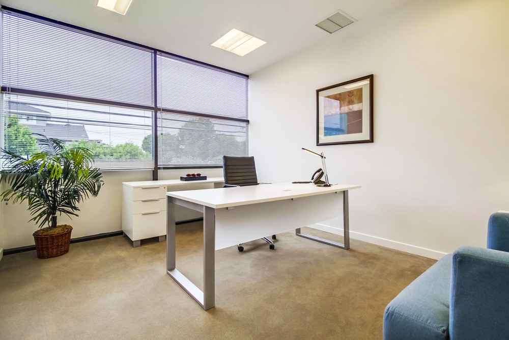Barrister Suites Office Lease A modern, minimalist office with a white desk, black chair, desk lamp, white drawer unit, potted plant, framed artwork on the wall, and large windows letting in natural light.
