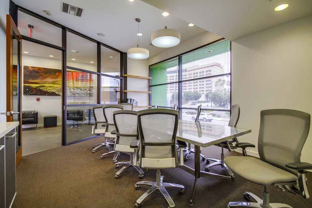 Barrister Suites Office Lease Modern office conference room with a long table, several mesh chairs, large windows with a city view, and glass walls separating the space from an adjacent lounge area.