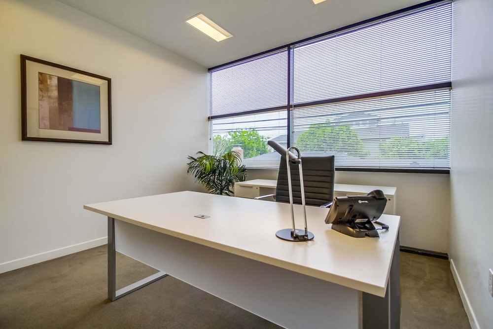 Barrister Suites Office Lease Modern office with a white desk, black chair, desk lamp, telephone, and a potted plant near large windows with blinds. Abstract art hangs on the wall, and trees are visible outside.